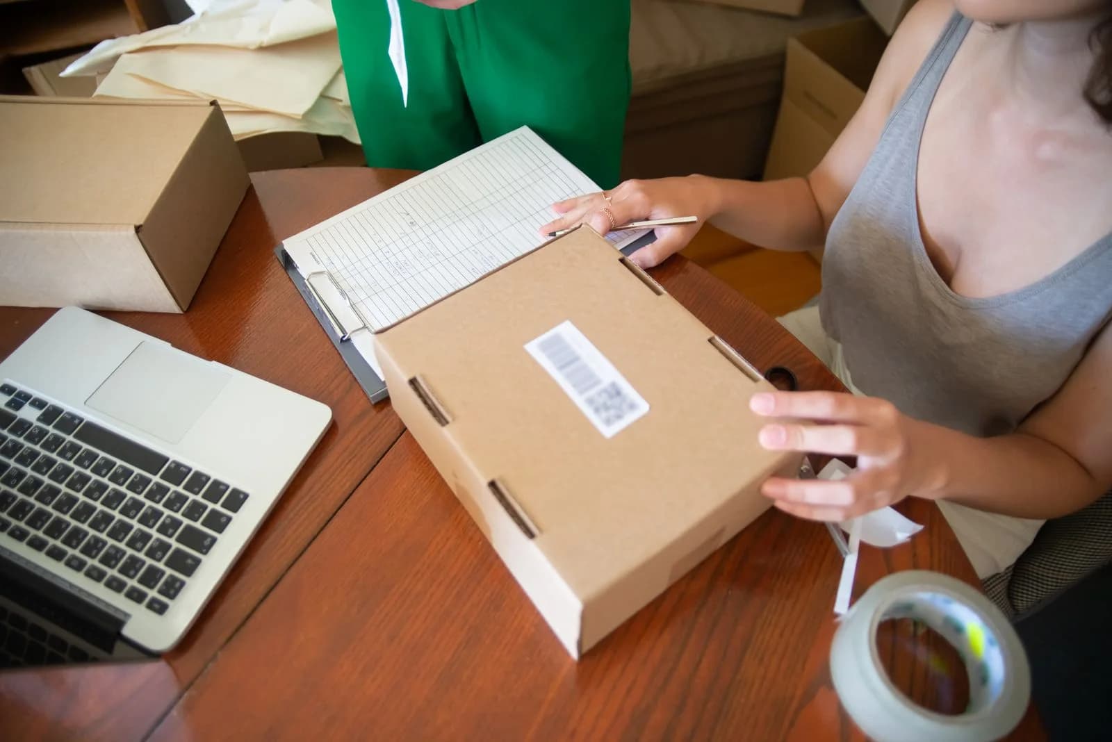 A person checking an order sheet while packing a parcel beside a laptop.